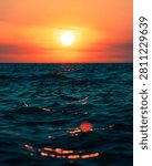 Dramatic sunset over the ocean with deep blue waves reflecting the vibrant orange and red sky. The shallow depth of field creates beautiful golden bokeh on the water surface. Captured in East Java, Indonesia.