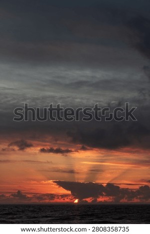 Dramatic sunset over the ocean with glowing orange sky, dark clouds, and calm waves on the horizon
