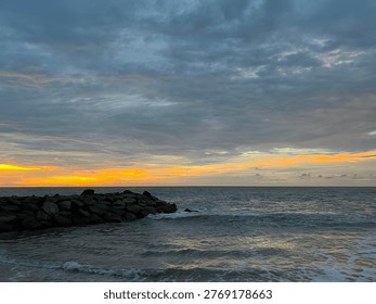 Dramatic sunset over the ocean with rocky shoreline and gentle waves under a cloudy sky