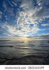 Dramatic sunset over the ocean with scattered clouds, golden sun rays shining on gentle waves, and silhouettes of people enjoying the beach landscape.