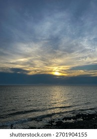 Dramatic sunset over the ocean, with the sun breaking through dark, moody clouds and reflecting on the sea water above a rocky shore.