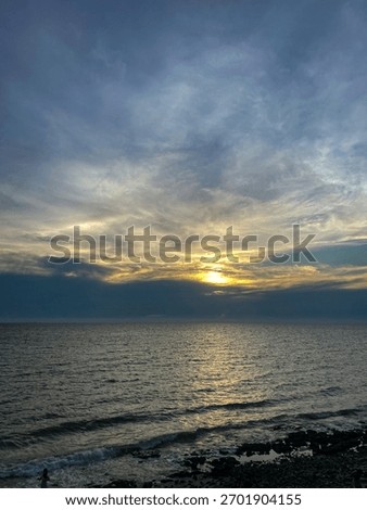 Dramatic sunset over the ocean, with the sun breaking through dark, moody clouds and reflecting on the sea water above a rocky shore.