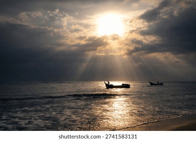 Dramatic sunset over the ocean with traditional fishing boats silhouetted against golden sun rays breaking through clouds, reflecting on calm sea water and sandy beach shoreline.