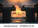 A dramatic sunset over the ocean viewed through a broken bridge. Waves crash below as a bird flies towards the vibrant sky.
