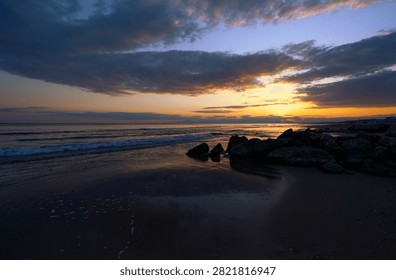 A dramatic sunset over a rocky beach with gentle ocean waves, reflective wet sand and brooding clouds casting warm orange and purple light across the twilight shoreline.