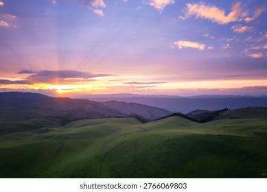 Dramatic sunset over the rolling green hills of Kala Jun grassland, with radiant sunbeams piercing through colorful clouds and distant mountain ranges.