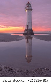 Dramatic sunset over a sandy beach with a tall lighthouse reflected in a calm tidal pool. Soft pink and orange skies add to the serene ambiance.