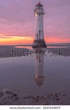 Dramatic sunset over a sandy beach with a tall lighthouse reflected in a calm tidal pool. Soft pink and orange skies add to the serene ambiance.