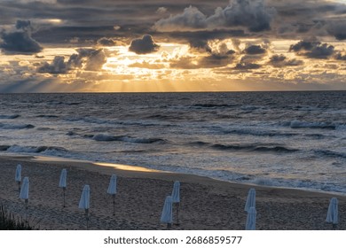 A dramatic sunset over the sea. Rays of sun break through dark clouds. Umbrellas are rolled up on the beach. Background. Landscape.