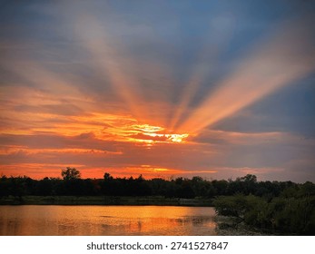 Dramatic sunset over a tranquil lake with radiant sunbeams streaming through vibrant orange and crimson clouds. Golden reflections shimmer across calm waters, framed by a lush tree line, creating a breathtaking evening landscape filled with warmth, serenity, and natural beauty. A striking scene of light, color, and peaceful outdoor scenery.