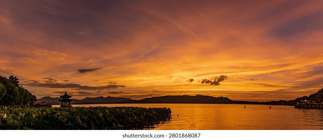Dramatic sunset over West Lake in Hangzhou, China, with a traditional pavilion, lotus leaves, calm water, and mountain silhouettes under a glowing orange sky.