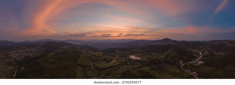 Dramatic sunset panorama over green rolling hills and valleys. Aerial view of a rural landscape with winding roads, a small lake, and a vibrant colorful sky. Wide background with copy space.