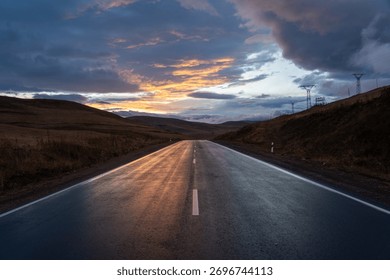 Dramatic Sunset Reflection on Wet Asphalt Road Leading Through Dry Hills Under Dark Clouds. Journey, Hope, and Power Lines Concept.