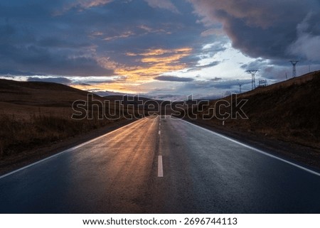 Dramatic Sunset Reflection on Wet Asphalt Road Leading Through Dry Hills Under Dark Clouds. Journey, Hope, and Power Lines Concept.
