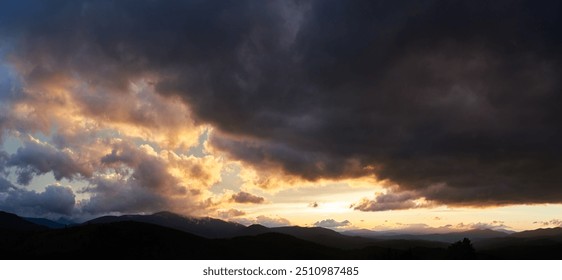 Dramatic sunset scene with dark, brooding clouds casting shadows over sky. Horizon glows with warm hues of orange and pink, contrasting against silhouettes of mountains, creating moody atmosphere.