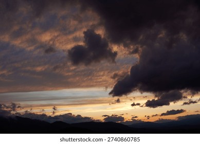 Dramatic sunset scene with dark, brooding clouds casting shadows over sky. Horizon glows with warm hues of orange and pink, contrasting against silhouettes of mountains, creating moody atmosphere.