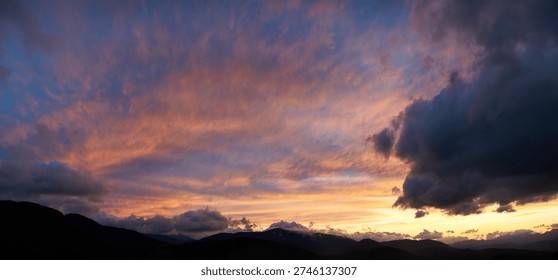 Dramatic sunset scene with dark, brooding clouds casting shadows over sky. Horizon glows with warm hues of orange and pink, contrasting against silhouettes of mountains, creating moody atmosphere.