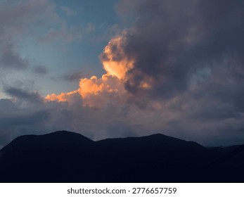 A dramatic sunset scene featuring dark, silhouetted mountain peaks against a dynamic sky. Large, billowing cumulus clouds are illuminated from within by a golden-orange glow from the setting sun, contrasting sharply with the deep blue and grey tones of the surrounding storm clouds. This atmospheric landscape captures a sense of majesty and natural power, making it ideal for cinematic backgrounds, weather-related articles, or inspirational travel content.