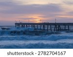 A dramatic sunset scene at Pacifica Municipal Pier in Pacifica, California, with powerful ocean waves crashing beneath the long concrete fishing pier.