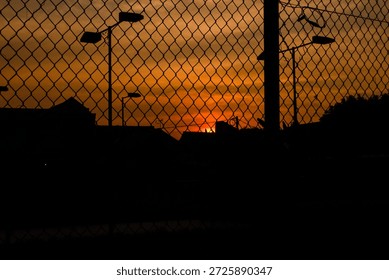 Dramatic sunset scene viewed through a chain link fence, with the sun setting on the horizon and casting warm orange and golden tones across the sky. Silhouettes of urban elements such as street light