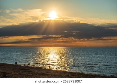 A dramatic sunset shines through dark clouds, casting golden light on the sea and a beach where few silhouettes of people walk along the shore. The scene is vibrant and twilight.