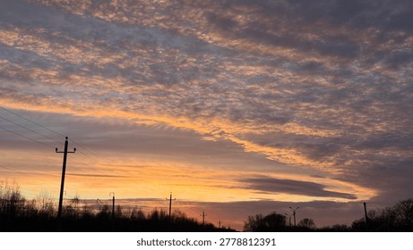 Dramatic sunset sky with bright orange and pink textured clouds stretching over the silhouettes of trees and power lines.