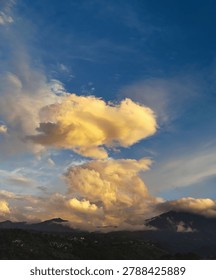 Dramatic sunset sky featuring massive golden-illuminated clouds against deep blue, towering over misty dark mountains and a lush green hillside village.