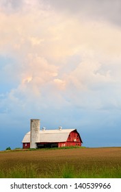 A dramatic sunset sky hangs over a red barn with silo and basketball hoop in the Midwestern United States.