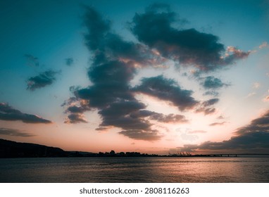 Dramatic sunset sky over calm waterfront with city skyline, bridge and cloud silhouettes, peaceful urban landscape for travel, relaxation and evening atmosphere concept