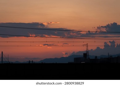 Dramatic sunset sky over distant mountains and city silhouette layered with electrical power lines and overhead wires, Japan.
