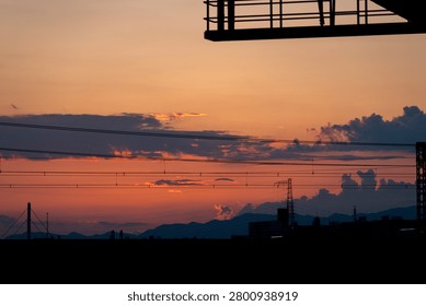 Dramatic sunset sky over distant mountains and city silhouette layered with electrical power lines and overhead wires, Japan.