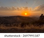 A dramatic sunset sky over rolling hills with layers of clouds illuminated in orange and gold tones. A breathtaking evening scene.