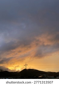 Dramatic sunset sky over a small town with dark storm clouds and golden sunlight breaking through, silhouettes of mountains and power lines in the countryside.