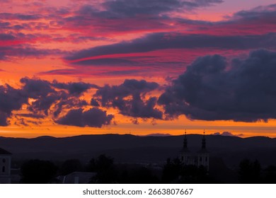 A dramatic sunset sky with vibrant orange and pink clouds hovering over a silhouette of hills and a historic church.