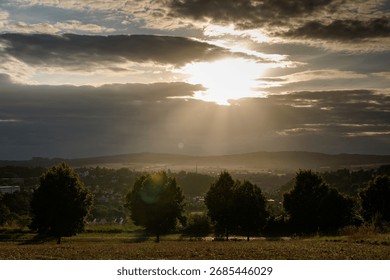 Dramatic sunset with sun rays breaking through thick clouds over a rural valley, illuminating the landscape with trees in the foreground and a town in the distance.