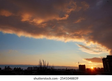 Dramatic sunset with vibrant clouds over a city skyline. The horizon glows with the setting sun, casting a golden light over the urban landscape and silhouetting buildings.