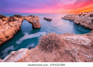 Dramatic sunset view of Arco de Albandeira at Praia de Albandeira in Algarve, Portugal, with pink sky, limestone sea arch, Atlantic water, and a high Atlantic view over the cliff edge.