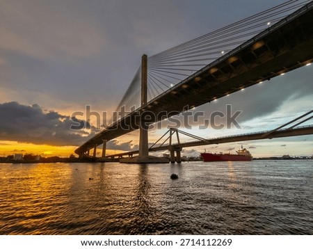 Dramatic sunset view from beneath the Pumarejo Bridge over the Magdalena River in Barranquilla, Colombia, highlighting bridge pillars, structural design, river reflections and transportation infrastructure.