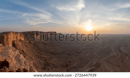 Dramatic sunset view from the Edge of the World near Riyadh, with sheer sandstone cliffs dropping into vast desert plains under streaked skies.