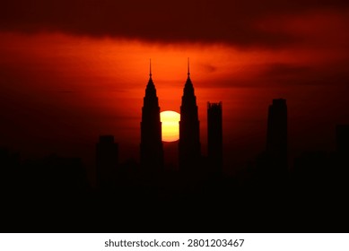 Dramatic sunset view of the Kuala Lumpur city skyline with the sun positioned between the iconic Petronas Twin Towers. Silhouette of modern skyscrapers against a vibrant red and orange evening sky in Malaysia.