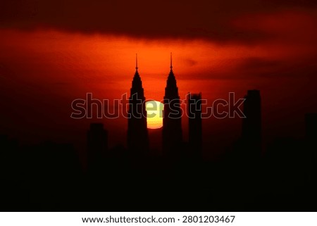 Dramatic sunset view of the Kuala Lumpur city skyline with the sun positioned between the iconic Petronas Twin Towers. Silhouette of modern skyscrapers against a vibrant red and orange evening sky in Malaysia.