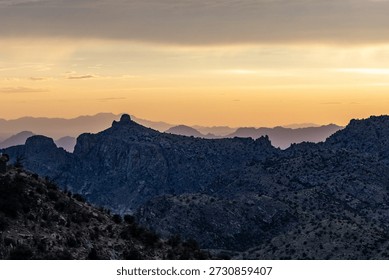 Dramatic sunset view from Mount Lemmon showing golden light on rugged rocky ridges layered formations distant mountain ranges and valley under cloudy sky
