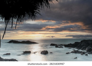 Dramatic sunset view over a rocky coast with long exposure soft waves, moody cloud formations, and silhouette palm foliage foreground. Serene tropical beach landscape.