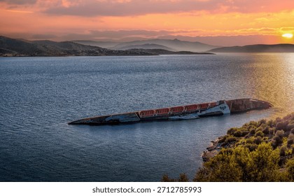 A dramatic sunset view of the sunken MS City of York, a former cruise ship, resting in the Gulf of Elefsina, Greece. The wreck is partially submerged in the calm waters, with the coastline and surrounding hills visible in the background.