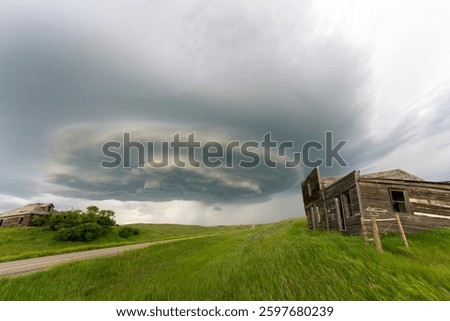 Dramatic supercell storm cloud forming over abandoned wooden houses and vast prairie fields in rural landscape