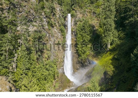 Dramatic tall waterfall plunges through lush evergreen forest with mist and a rainbow in British Columbia, Canada.