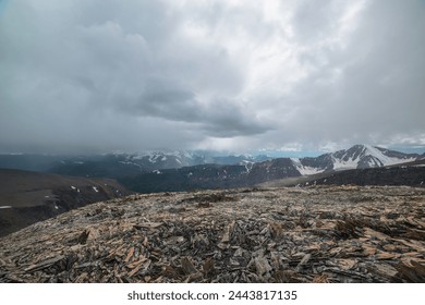 Dramatic top view from high stony pass to alpine valley against large mountain range with snow-capped peaks in gray rainy low clouds. Beautiful big snowy mountain tops in rain under grey cloudy sky.