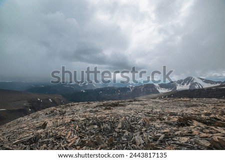 Dramatic top view from high stony pass to alpine valley against large mountain range with snow-capped peaks in gray rainy low clouds. Beautiful big snowy mountain tops in rain under grey cloudy sky.