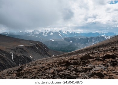 Dramatic top view from stone pass above alpine valley to large mountain range with snow-capped peaks on horizon in gray cloudy sky. Rocky steep slope and high snowy tops far away under rainy clouds.
