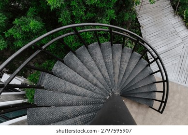 A dramatic top-down, high-angle shot looking down a black metal spiral staircase with diamond-plate textured steps. The circular geometry of the staircase contrasts sharply with the background below, which shows a light-colored wooden plank walkway, concrete ground, and dense green tropical foliage.